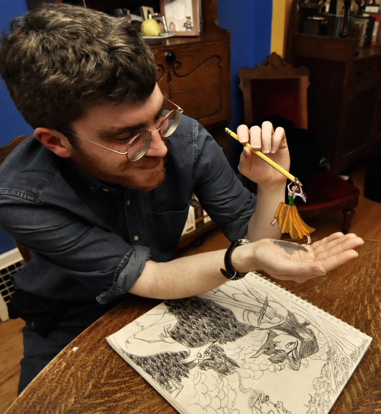 Picture of Tristan, a young, white male with glasses, next to some of his art and holding a pencil with one of his characters photoshopped to look like it is holding onto the end.