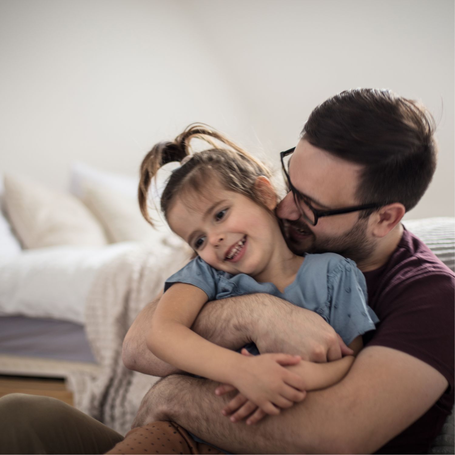 Photo of a father and daughter hugging and smiling while sitting next to a bed in their home