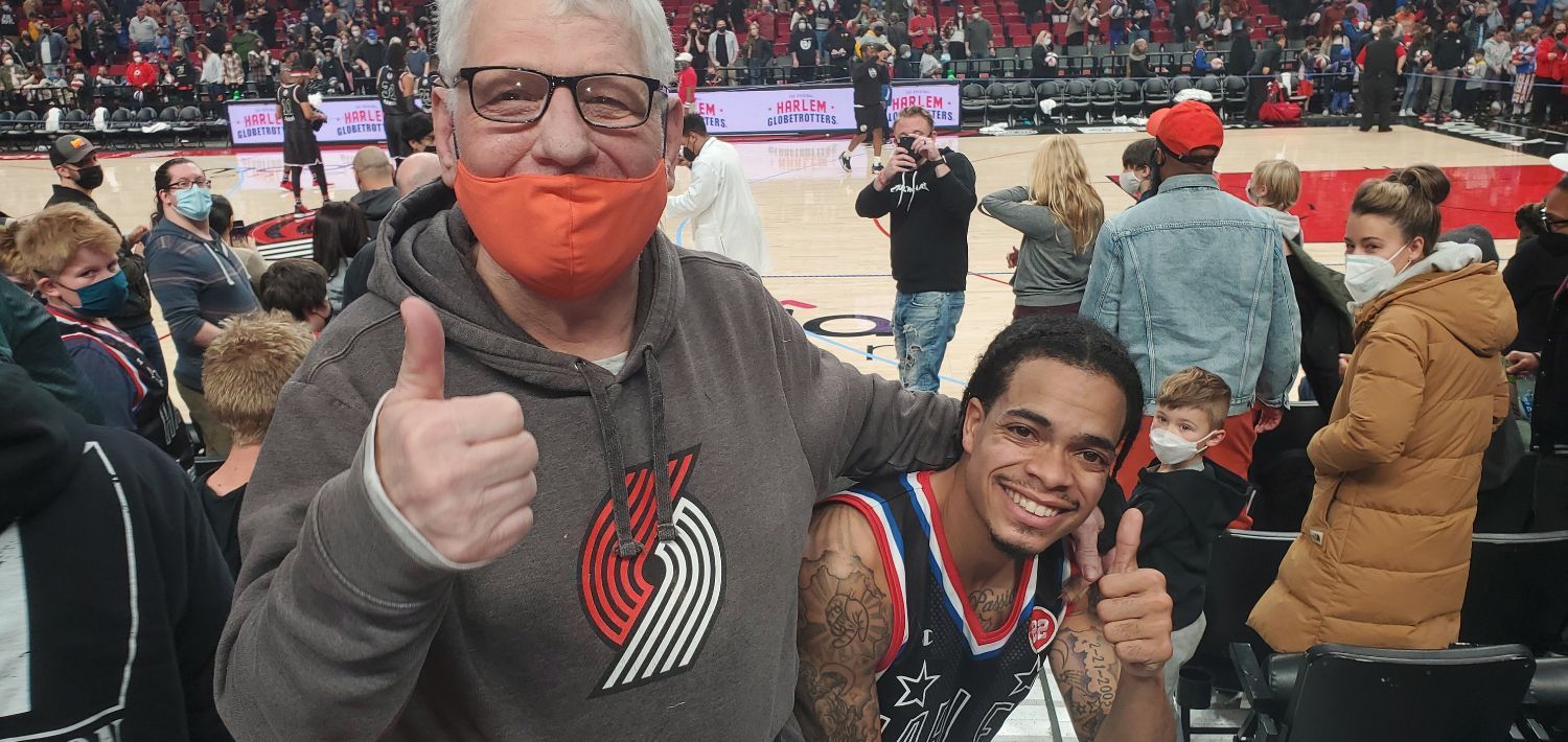 Picture of a supported individual, Steve, an older white man with glasses, and a member of the Harlem Globetrotters, a young black man with cornrows with a part in the middle, at a basketball game. Both are smiling and giving the thumbs up.