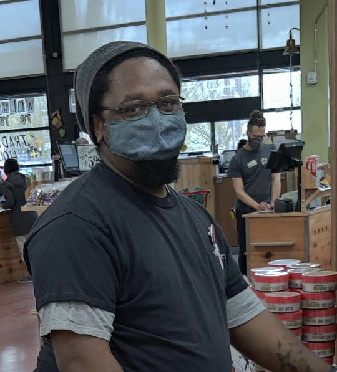 Jonathan, a young black man in a navy blue t-shirt, with a light blue mask, and a light gray beanie on stands smiling in Trader Joe's.
