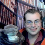 Devon, a young white man with brown hair and glasses smiles while he holds a hippo puppet.