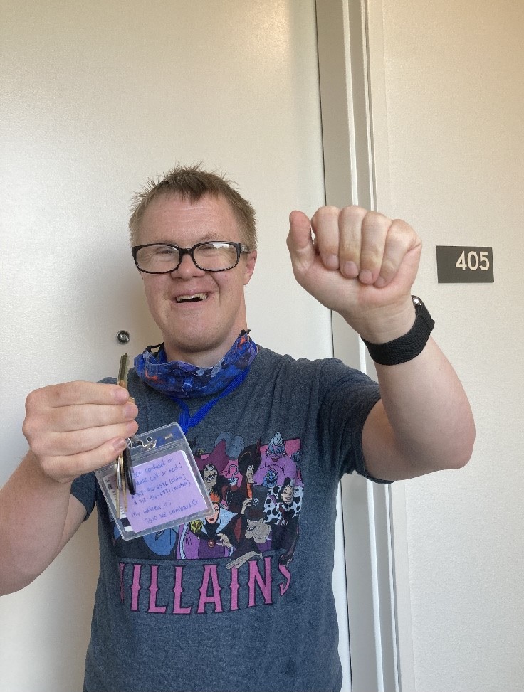 Matt, a young white man with dirty blonde hair and glasses, smiles with one fist in the air and an other one holding an apartment key.