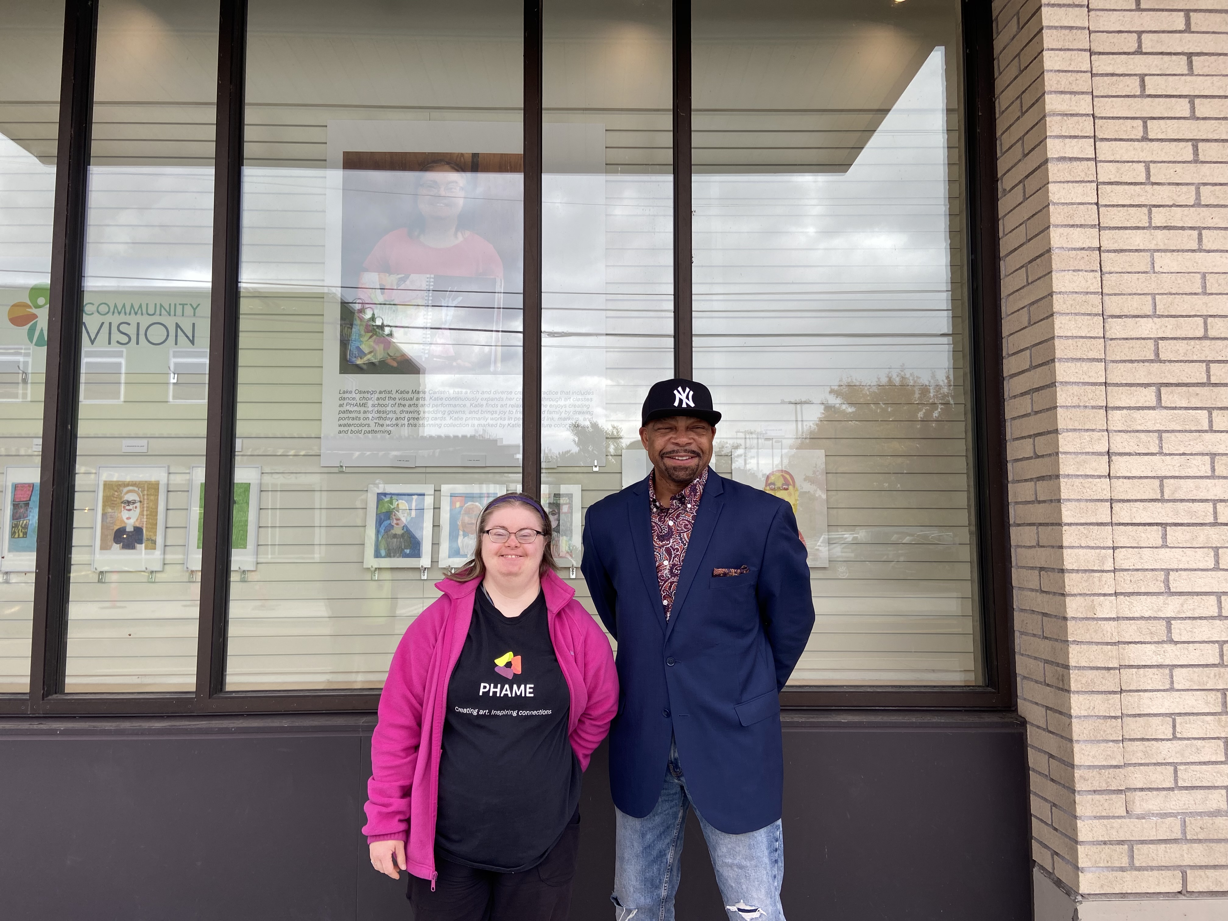 Katie Carlsen, a young white woman in a pink jacket, and John Canda, Community Engagement Director, a black man in a blue blazer and black hat, stand in front of Katie's exhibition.