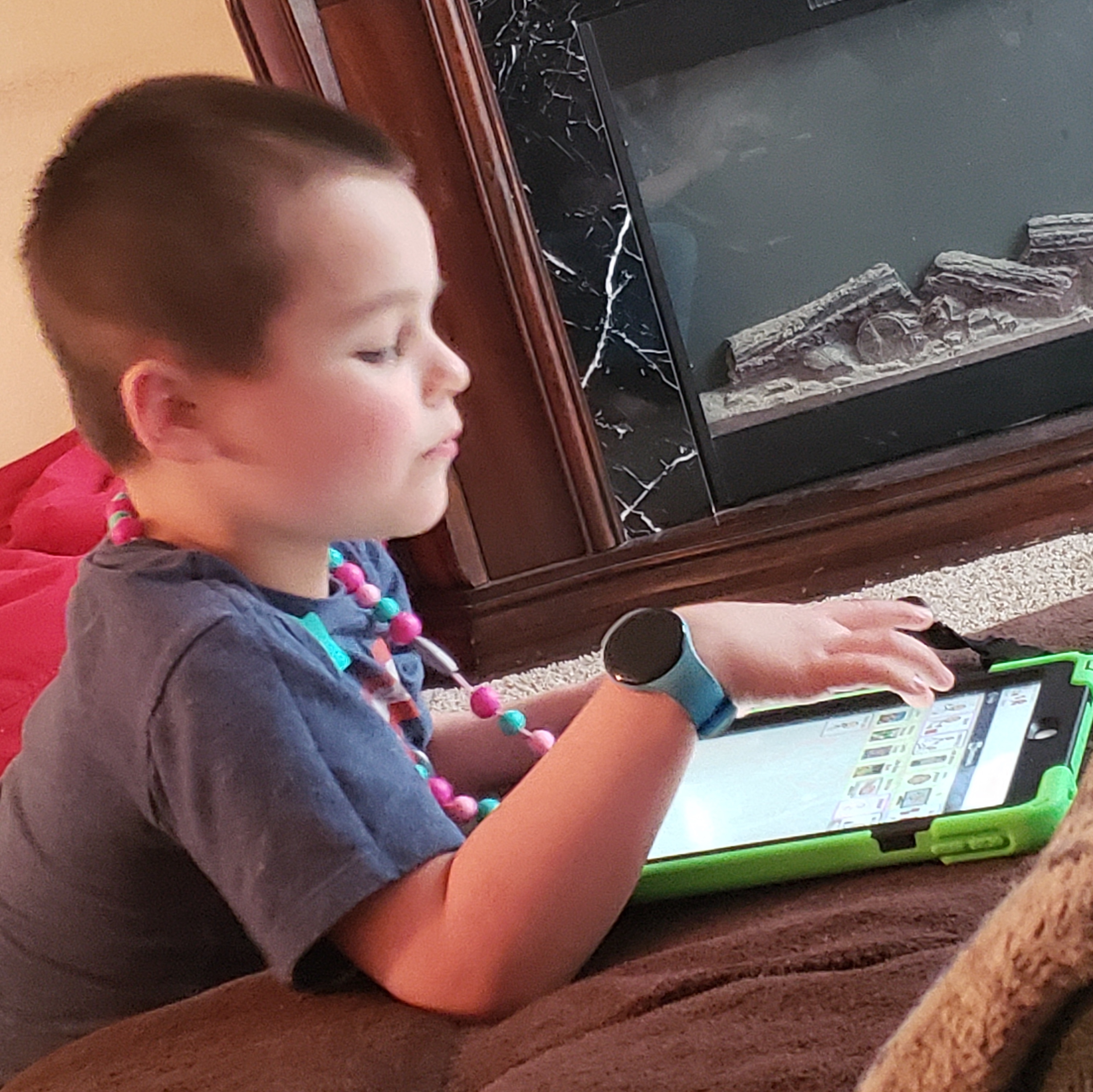 Kaysen, a young boy with short brown hair, a gray shirt, and rainbow beaded necklace, using an iPad.